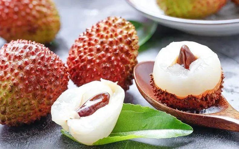 一颗被横向剖开的妃子笑荔枝,展示其半透明晶莹的乳白色果肉,果肉包裹着一个中等大小的褐色种子,果皮外侧为红绿相间纹理,内侧为白色海绵层,背景为木质砧板,突出食物的自然质感 妃子笑荔枝切开展示果肉与果核