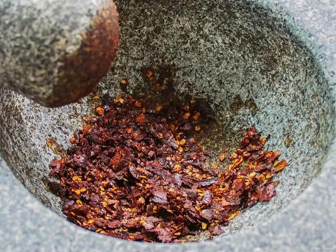 石臼中正在用木杵捣碎烤好的线椒和蒜末,已有部分成泥状,边缘可见黑色焦皮碎片,背景可见切好的茄子条和皮蛋块 石臼中正在用木杵捣碎烤好的线椒和蒜末,已有部分成泥状,边缘可见黑色焦皮碎片