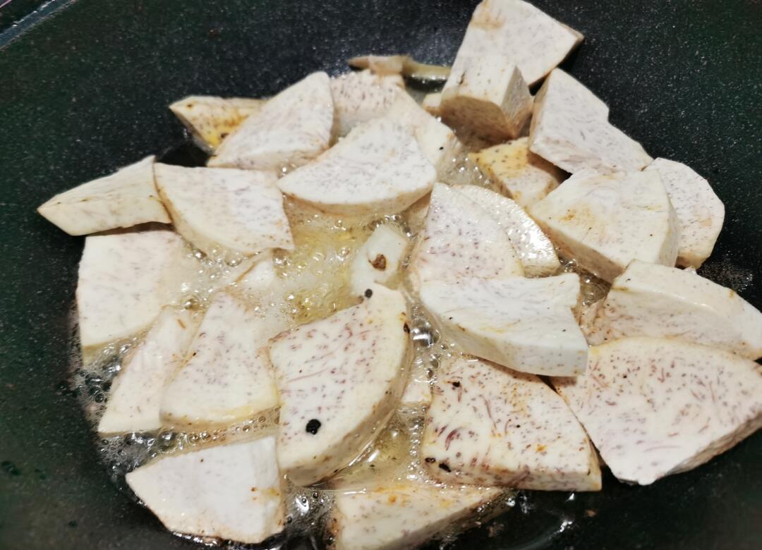 平底锅中正在油炸的浅黄色芋头片,油面轻微冒泡,芋头边缘呈现微微金黄焦边状态,旁边放置一双木筷用于翻动,背景可见厨房操作台和调料瓶 芋头油炸过程图