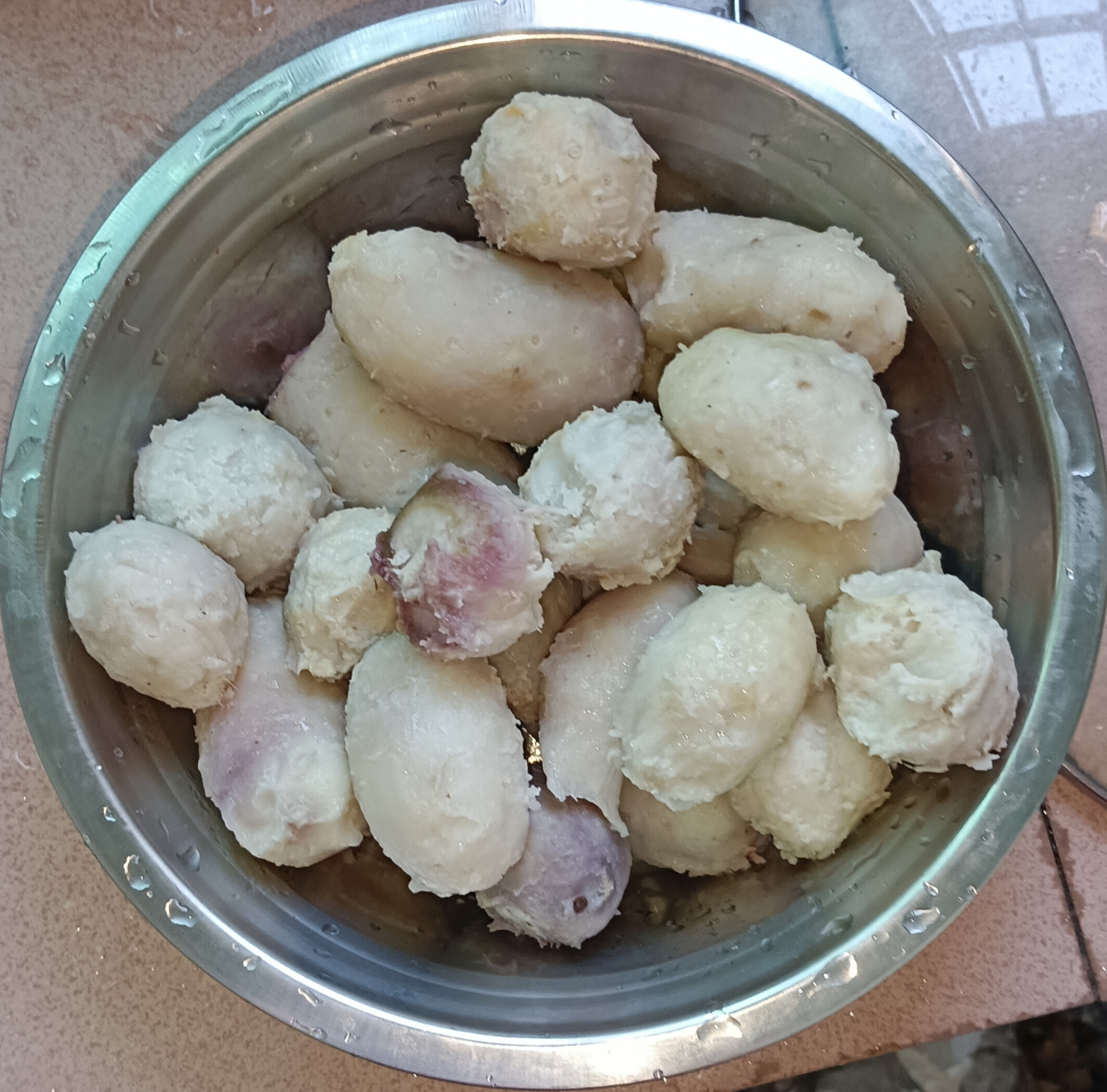 一位厨师正在不锈钢碗中用木勺搅拌刚蒸好的芋头,碗中为细腻的灰白色芋泥,灶台上放有椰浆罐、黄油块和量勺,背景为整洁的开放式厨房,窗外透进自然光 厨房中制作芋泥的过程