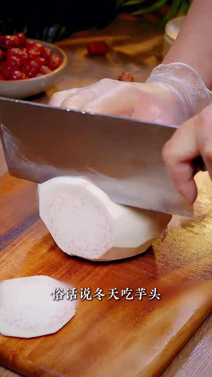 一位厨师戴着橡胶手套正在用刀给一个表皮粗糙的毛芋头削皮,案板上有少量白色黏液,旁边放着一碗清水和一把备用刀具,背景为干净整洁的厨房环境 毛芋头去皮过程特写