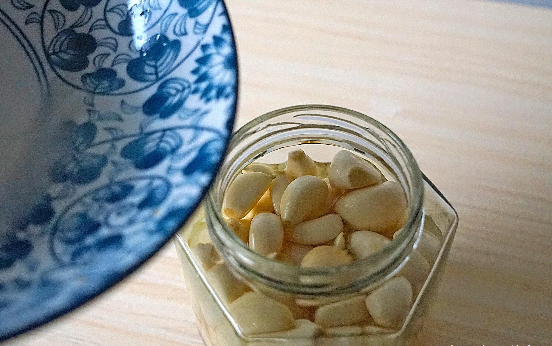 一个透明玻璃蒜罐内部特写,展示浸泡在淡黄色发酵液中的乳白色蒜米,部分蒜瓣边缘呈现轻微琥珀色,罐壁附着细小气泡,背景为木质厨房台面,体现家庭自然发酵场景 发酵过程示意图
