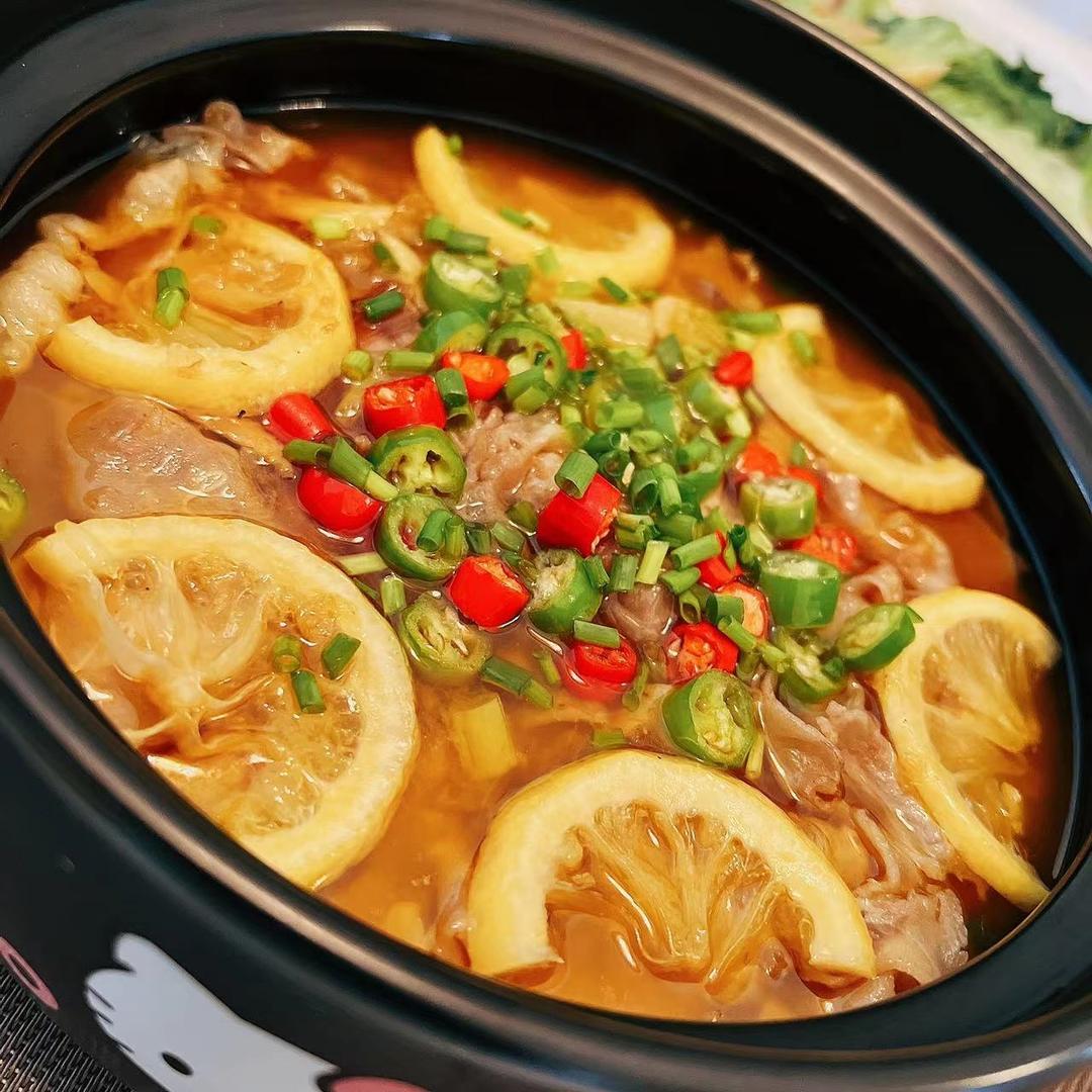 一锅正在炖煮中的柠檬泡椒金汤,汤色呈明亮的金黄色,表面漂浮着红亮的泡椒段和淡黄色柠檬片,热气升腾,旁边摆放着新鲜泡椒、柠檬和姜蒜等原材料 图片描述