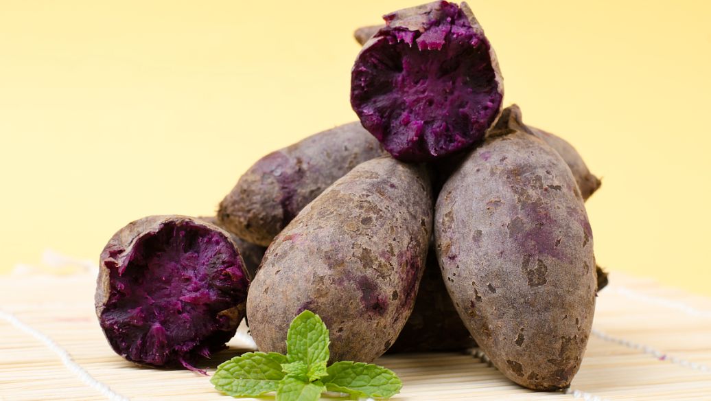 一个刚被横向切开的紫薯,露出鲜艳的深紫色果肉,切面湿润有光泽,周围摆放着几片完整的紫薯片和一根绿色的紫薯藤,背景为木质餐桌,突出食材的新鲜与自然美感 切开的紫薯特写展示紫色内芯