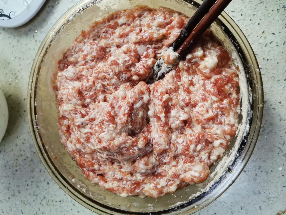 一个白色瓷碗中盛有混合中的香芹猪肉馅,筷子正顺着同一方向搅拌,馅料呈现出光泽细腻的状态,旁边摆放着酱油瓶、香油瓶和切碎的香芹 香芹猪肉馅搅拌过程