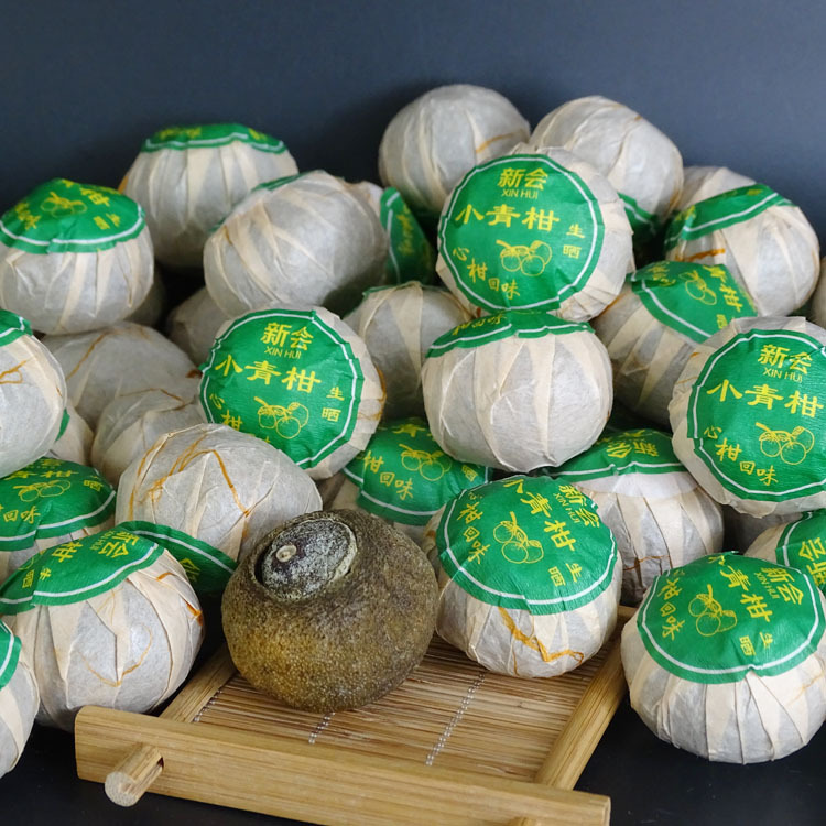 高清微距镜头拍摄的小青柑成品特写:直径约3.5cm的完整青柑果体,果皮厚约2.8mm,呈青绿至墨绿渐变色,表面可见密集凸起的透明油腺点(挥发油囊),顶部保留新鲜果蒂,内部熟普茶料隐约可见深褐色条索轮廓;摆放于天然竹编茶盘,背景为泛黄木纹老茶仓墙面,体现新会原产地与传统工艺语境 小青柑特写:青绿色未成熟柑果完整包裹紧压熟普茶球,表皮布满细密油胞,果蒂鲜绿,置于竹编茶盘上,背景虚化呈现岭南老茶仓木质纹理