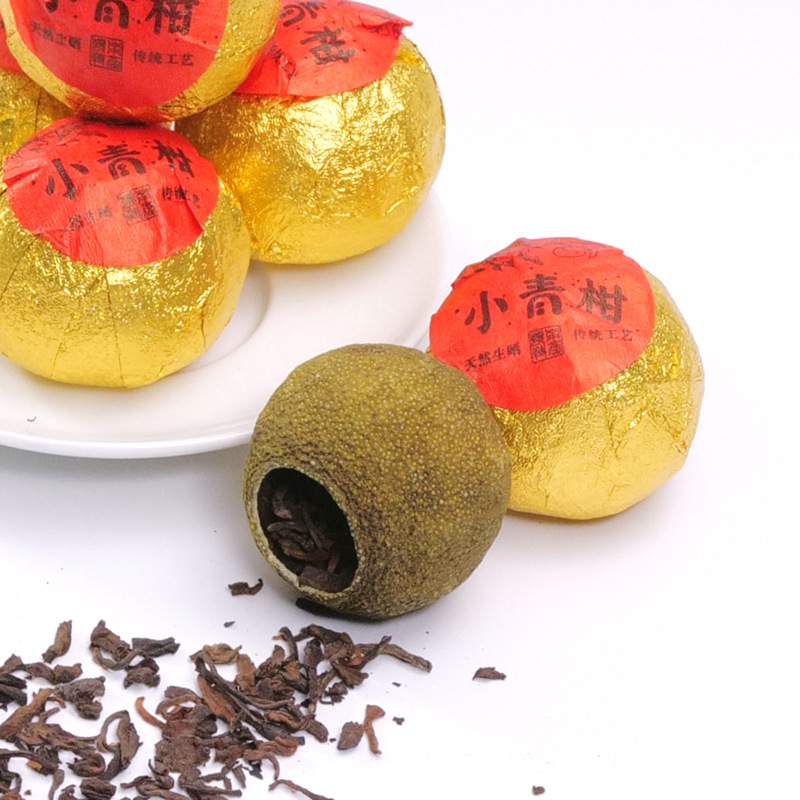 一颗完整的小青柑茶,表面呈深绿色,带有自然油斑,柑皮完整,旁边散落着少量剥开的茶叶,背景为木质茶盘,营造出传统茶艺氛围 小青柑特写