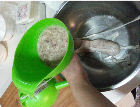 一名厨师正在使用不锈钢灌肠器将粉红色肉馅缓缓注入透明猪肠衣中,旁边放有扎线、针具和已完成的红肠串,背景为传统熏房木架 手工灌制哈尔滨蒜味红肠过程