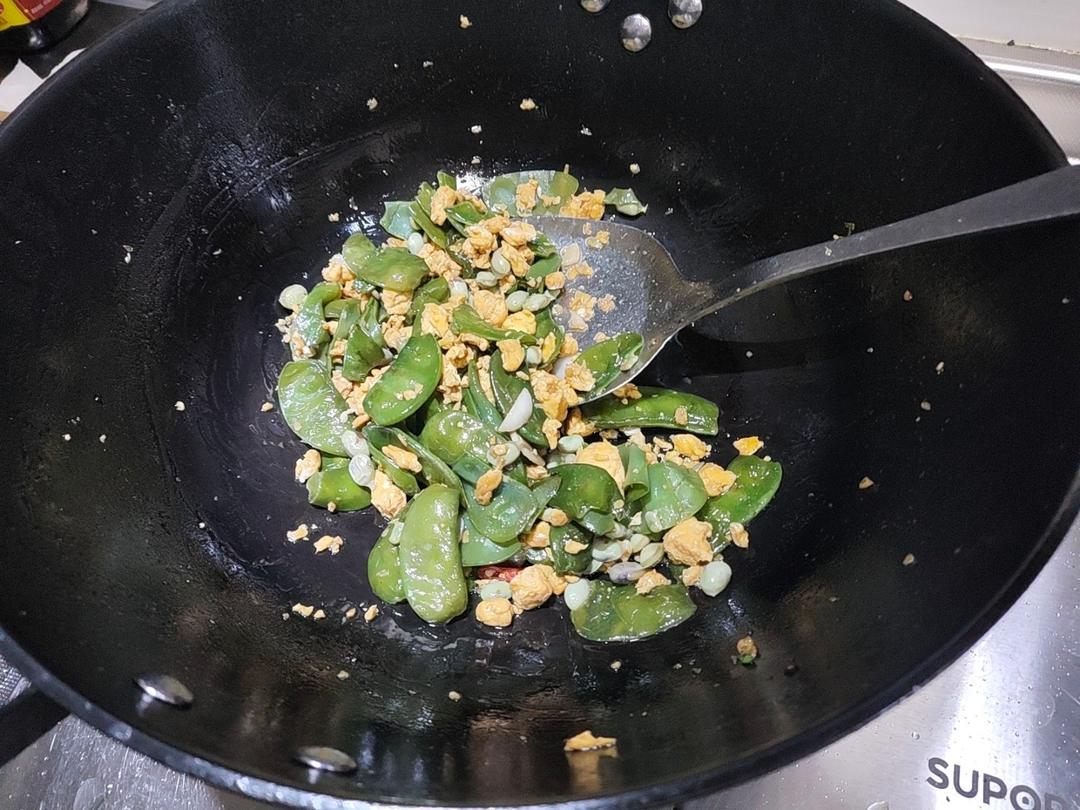 专业美食摄影:中式厨房环境,焦糖色铁锅内蚕豆与辅料动态翻炒瞬间,呈现高温快炒产生的‘锅气’视觉特征,豆粒形态饱满无焦糊,佐证科学火候控制 厨房场景:不锈钢炒锅中正在翻炒蚕豆,青绿色豆粒与淡黄色蒜末、红椒丝同炒,锅气升腾,油星细密,体现‘旺火快炒’传统技法