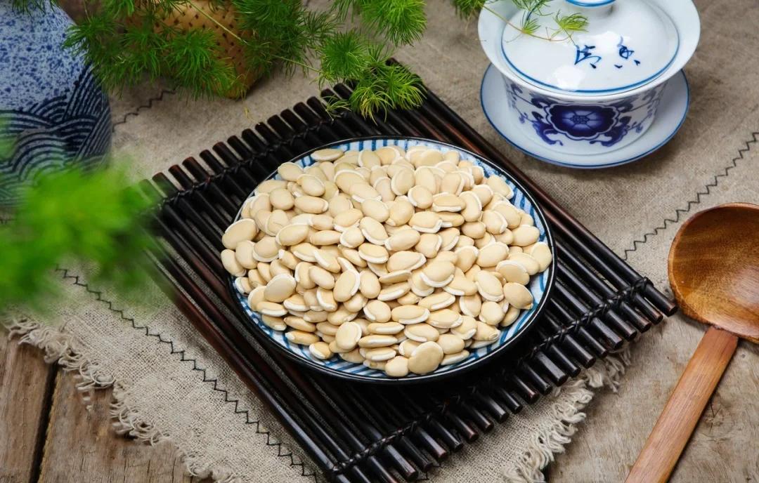 木质托盘上摆放着干燥的白扁豆种子与晒干的白扁豆花,旁边配有瓷杯中冲泡的花茶,背景为竹编篮子和中药材布袋,整体呈现传统中医药养生氛围 白扁豆与白扁豆花组合展示图