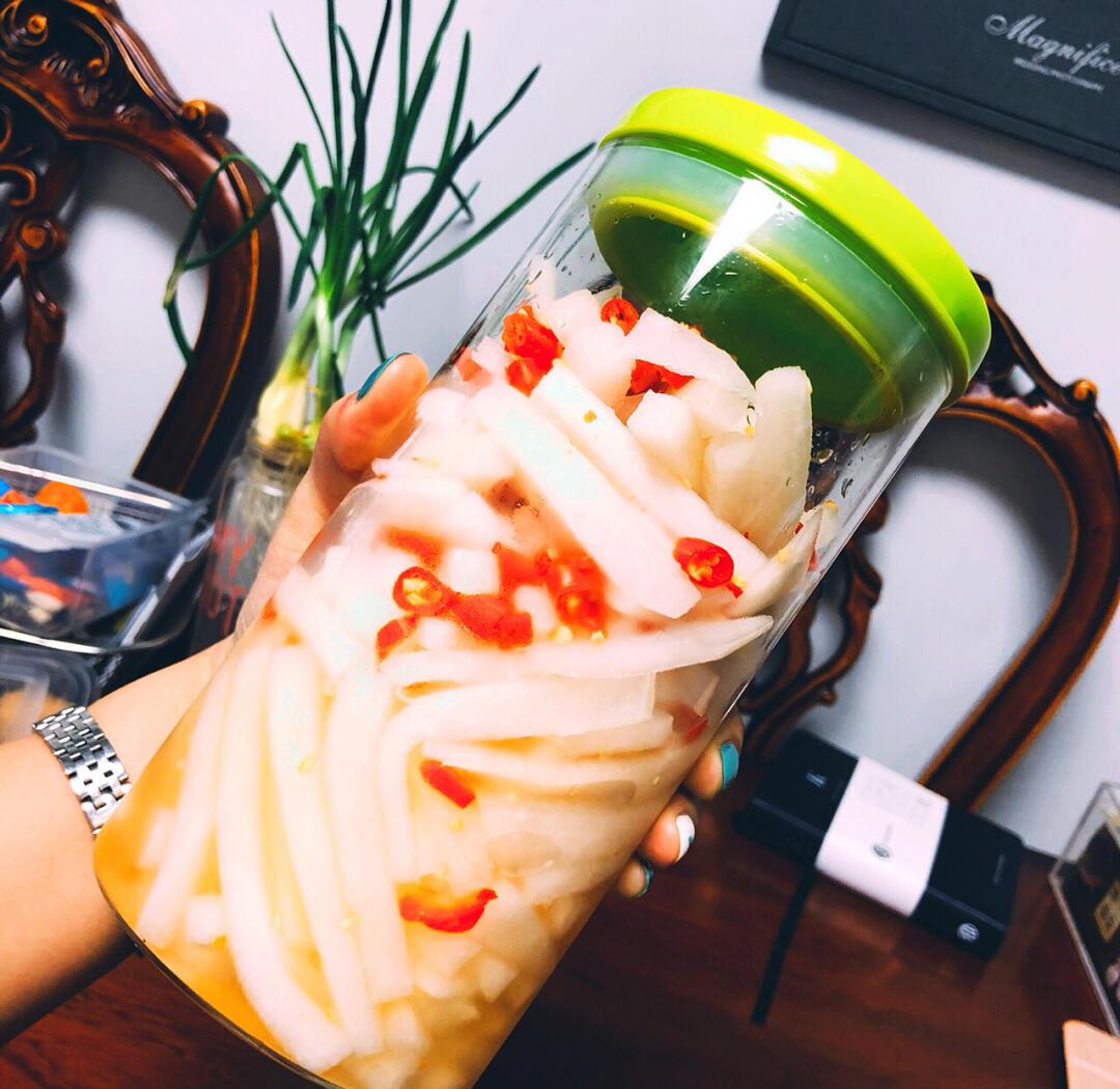 一个透明玻璃密封罐中装有紫红色的红皮萝卜块,表面覆盖着红亮的辣椒腌料,旁边摆放着切好的萝卜条、辣椒和蒜泥,背景为厨房操作台 红皮萝卜泡菜装罐过程