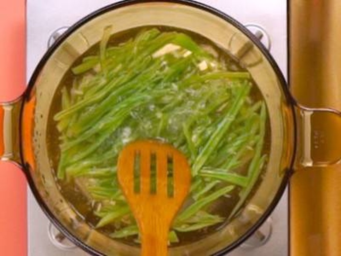 一位家庭主妇正在灶台前用铁锅翻炒芸豆角，锅中火焰升腾，食材呈翠绿色，蒜末清晰可见