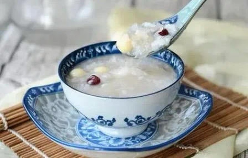 青花瓷碗中盛满乳白色的莲子山药芡实粥,表面微微冒热气,粥体浓稠可见完整的莲子和山药丁,旁边配有一双木筷和竹制餐巾盒,整体呈现温暖家常氛围 三宝粥成品图