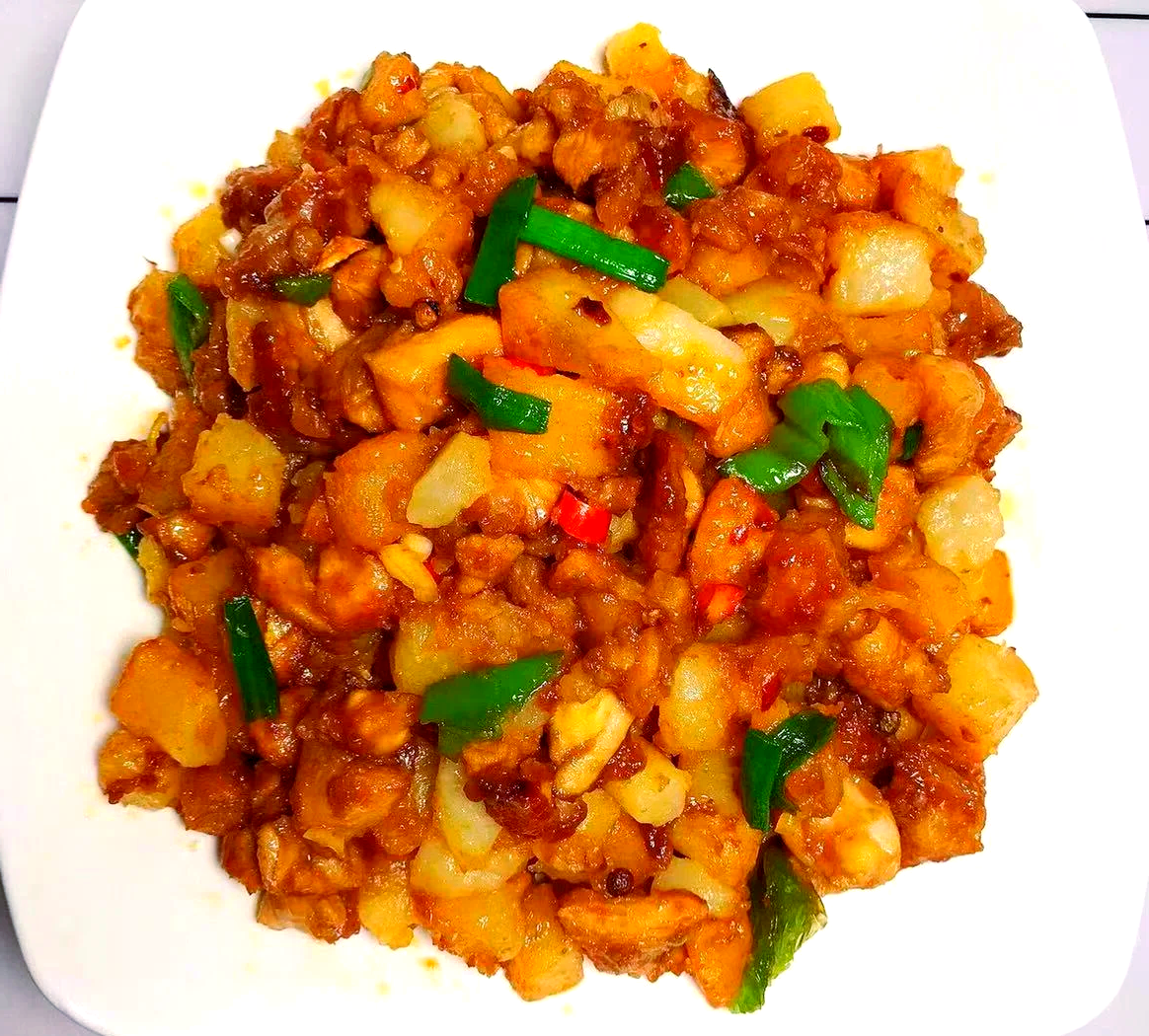一盘刚出锅的鸡丁炒土豆,金黄的土豆丁与乳白色的鸡丁交错,点缀绿色葱花和红色辣椒,盛放在白色瓷盘中,背景虚化的餐桌上摆有米饭和筷子 成品鸡丁炒土豆装盘效果