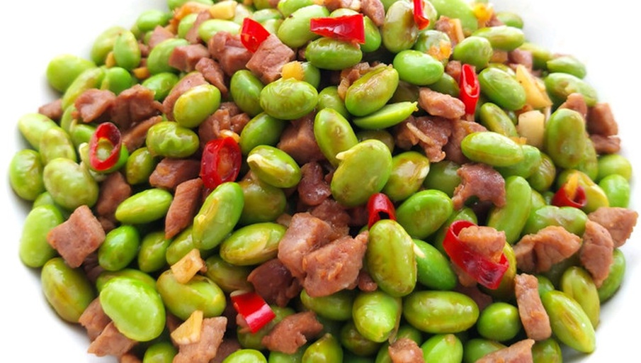 牛肉炒毛豆家常烹饪过程特写,突出食材本色对比与热力激发的天然香气,体现健康烹饪方式 家常烹饪场景:不锈钢炒锅中牛肉丁与毛豆粒同炒,色泽分明——酱色牛肉油亮,毛豆碧绿鲜亮,散落几粒红椒丁提色,锅气升腾,木质砧板旁放着刚剥好的毛豆和切好的牛肉