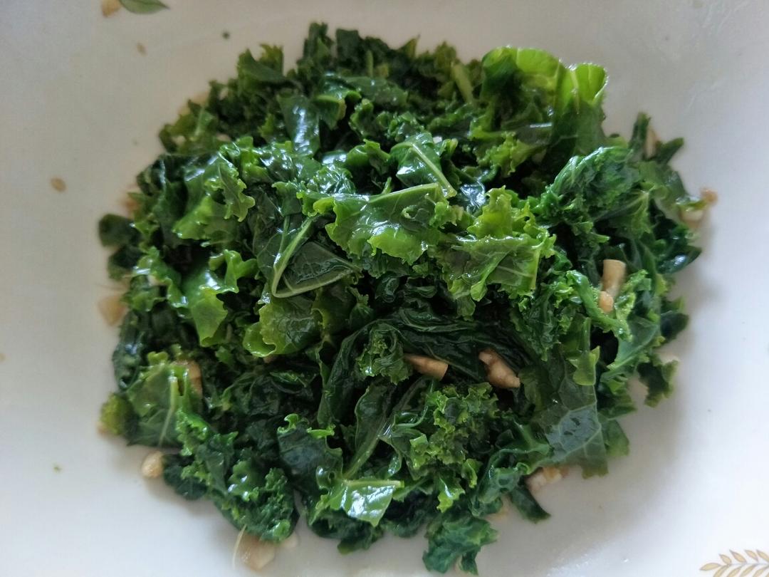一盘用橄榄油清炒的羽衣甘蓝菜肴,搭配蒜片和柠檬角,盛放在白色瓷盘中,背景虚化,突出食物色泽与质感 羽衣甘蓝料理成品图