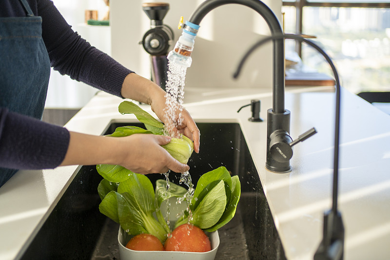 一位家庭主妇在厨房水槽边清洗一把新鲜油菜,水流清澈,油菜色泽鲜亮,旁边放着砧板和刀具,环境整洁温馨 家庭厨房中正在清洗油菜的过程