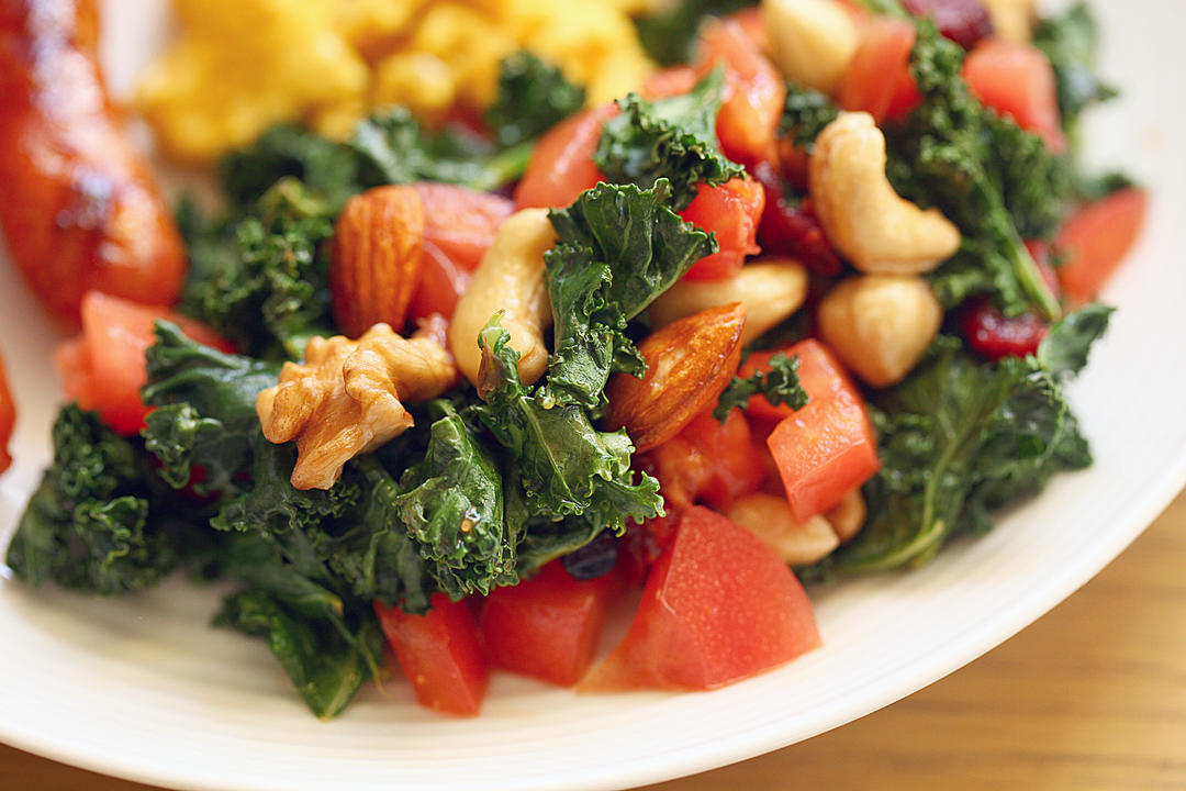 一碗色彩丰富的羽衣甘蓝茎丝沙拉,配有樱桃番茄、坚果和橄榄油调味,旁边放置一把木勺,背景为自然光线下的厨房台面 羽衣甘蓝茎制成的菜肴成品图