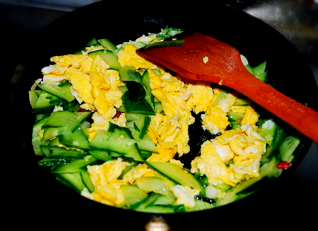平底炒锅中正在翻炒小黄瓜与鸡蛋，锅内食材呈现金黄色与翠绿色交织状态，灶火旺盛
