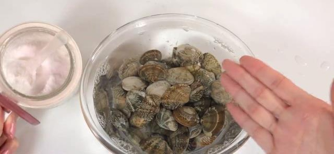 一盆清水中的花蛤正在浸泡吐沙,水中漂浮少量细沙,旁边放有食盐和香油瓶,背景为厨房操作台 花蛤吐沙处理过程