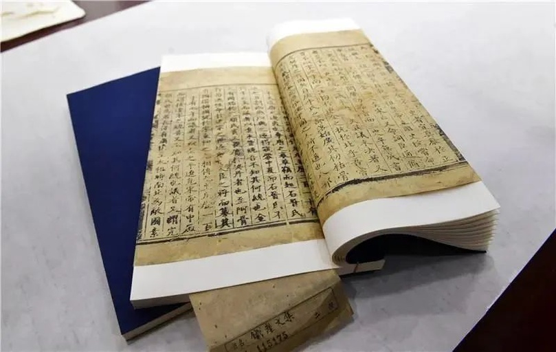 一本打开的羊皮纸古籍,页面上绘有精细的植物线描图,左侧为文字注释,使用古希腊文书写,右侧图画清晰呈现一种叶片层层包裹成球状的植物,旁边标注‘Krambe’或类似名称,周围散落着墨水瓶、羽毛笔和放大镜,体现学术研究氛围 古代手稿插图展示古希腊学者记录植物形态