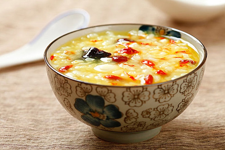 一碗盛好的小米枸杞粥特写，金黄粥体中点缀着红色枸杞，表面微微油亮，配以竹筷和原木托盘背景