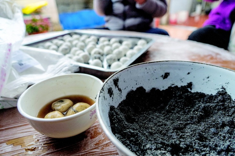 一位中年女性正在厨房用小火煮锅中的黑芝麻汤圆，锅边放置生姜片和红糖