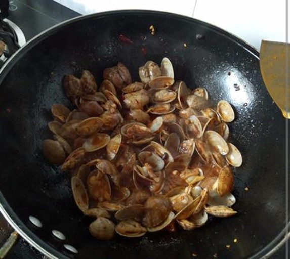 一位厨师正在家用铁锅高温翻炒花蛤,锅中花蛤已部分开口,伴有大量蒸汽和红色辣椒、蒜瓣、葱段等配料,锅边摆放着切好的青椒和调料瓶,背景为整洁的现代厨房 家庭厨房中正在铁锅内翻炒花蛤的场景