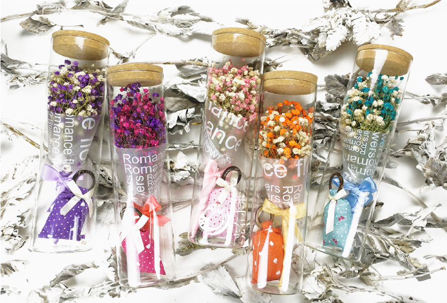 一张木质桌面上摆放着由‘和平’月季制作的透明永生花玻璃罩礼盒、浅粉色干花瓣罐装产品以及一瓶标有‘Rosa ‘Peace’ Essential Oil’的精油瓶,背景为柔和灯光下的花艺工作室环境 ‘和平’月季制成的永生花礼盒与精油产品展示