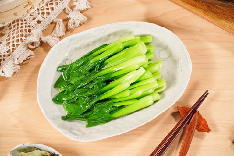 一位家庭主妇在厨房操作台前将新鲜油菜放入沸水中焯烫,旁边放着漏勺和冷水盆,用于后续过凉处理,灶台上可见干净整洁的厨具,体现健康烹饪理念 家庭厨房中正在焯水处理油菜的场景