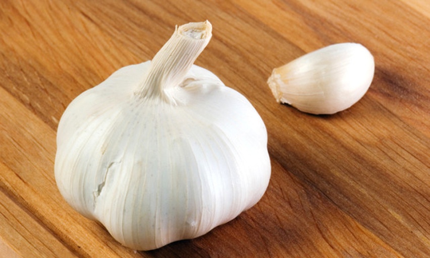 一个完整的大蒜头被切开展示内部结构,外层包裹着白色或淡紫色的膜状鳞片,内部清晰可见多个饱满的蒜瓣呈环形排列,中心为硬质的花茎基部。背景为木质砧板,旁边有一把小刀辅助展示 蒜头结构解剖图