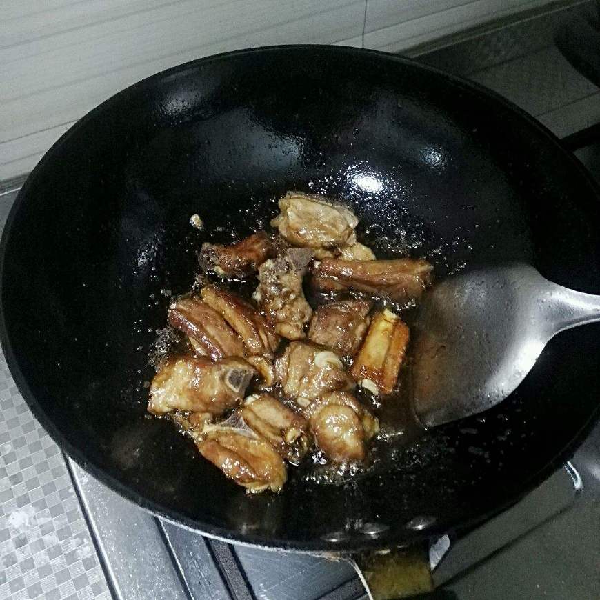 一口黑色铸铁炖锅内正在翻炒排骨与胡萝卜,姜蒜清晰可见,锅边有轻微油烟升起,炉火为中等蓝色火焰,展现家庭厨房真实烹饪场景 炖锅中翻炒食材过程