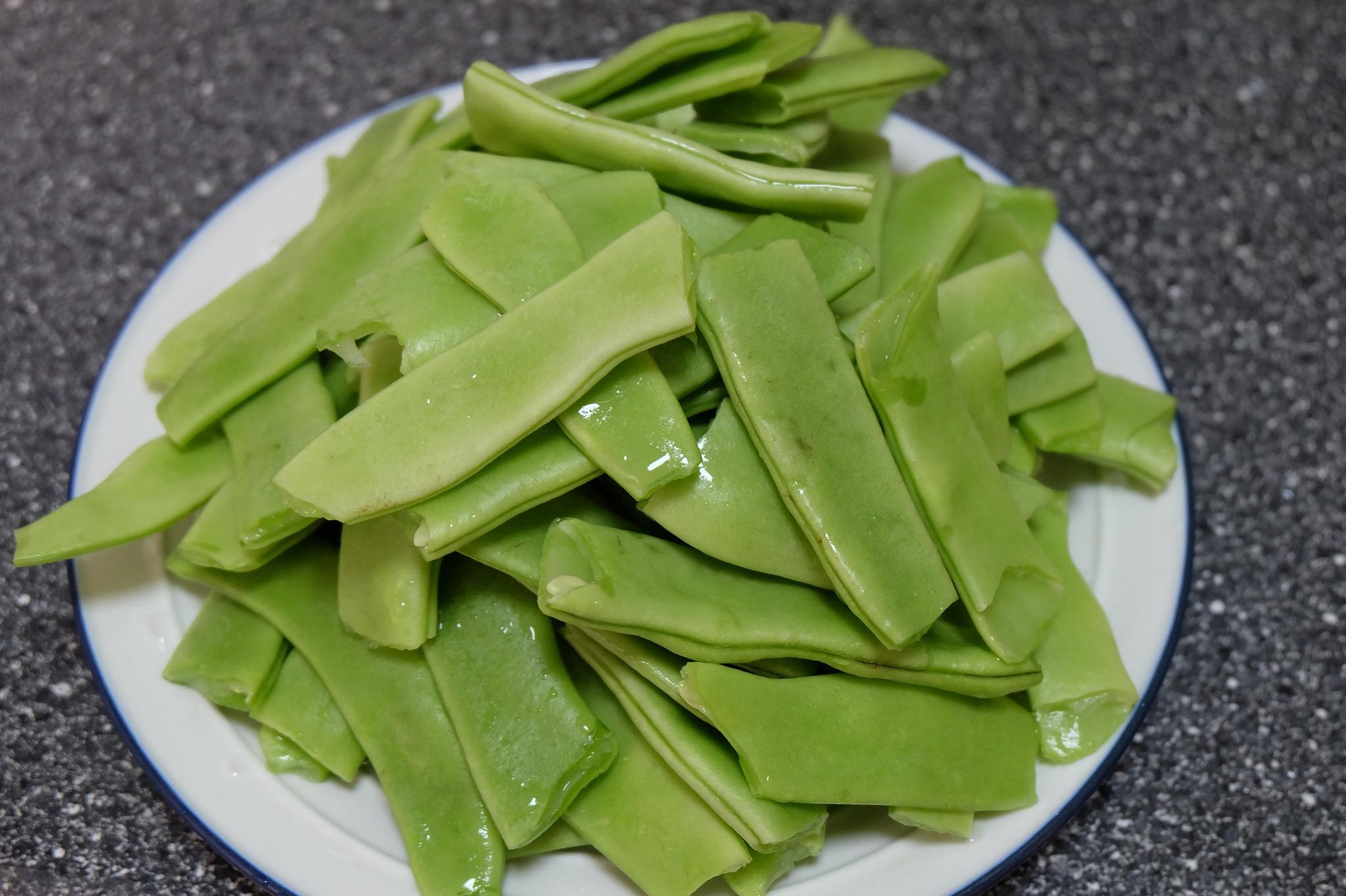 一位厨师正在灶台前用铁锅翻炒紫色扁豆,锅中扁豆呈半熟状态,部分已开始由紫转绿,旁边放有切好的蒜片和调料,厨房环境整洁明亮 厨房中正在翻炒紫扁豆的场景