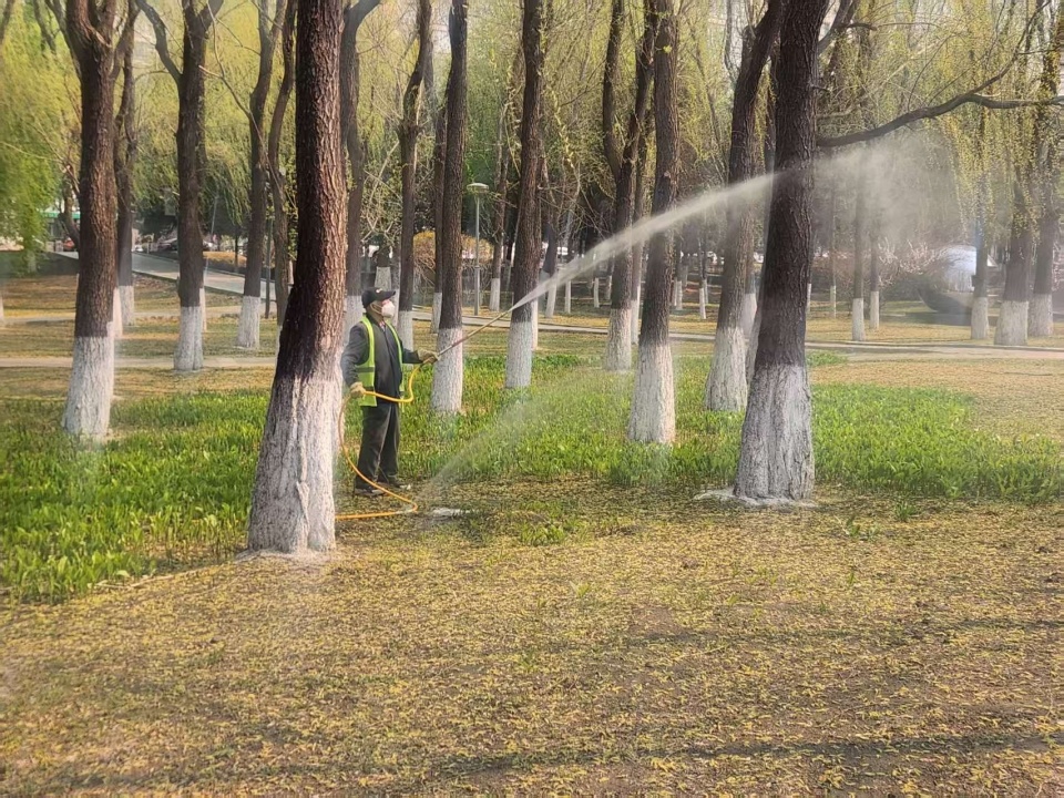 一名身穿防护服的工作人员正在公路护坡下方使用背负式电动喷雾器对成片杂草进行定向喷洒作业,喷头距杂草约30厘米,背景可见警示标志和行驶车辆 专业人员在公路边使用背负式喷雾器喷洒除草剂