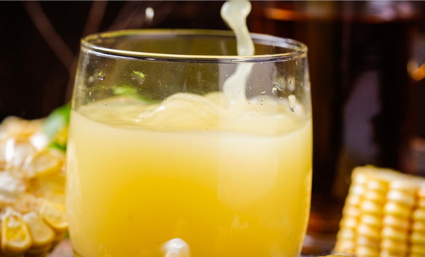 特写摄影:刚制成的熟玉米汁,色泽透亮无沉淀,泡沫细腻持久,直观呈现优质蒸制工艺带来的高溶解性与稳定性 新鲜玉米汁成品:琥珀色液体盛于透明玻璃杯中,表面有细腻泡沫,杯壁挂壁均匀,背景为竹蒸笼与黄糯玉米穗