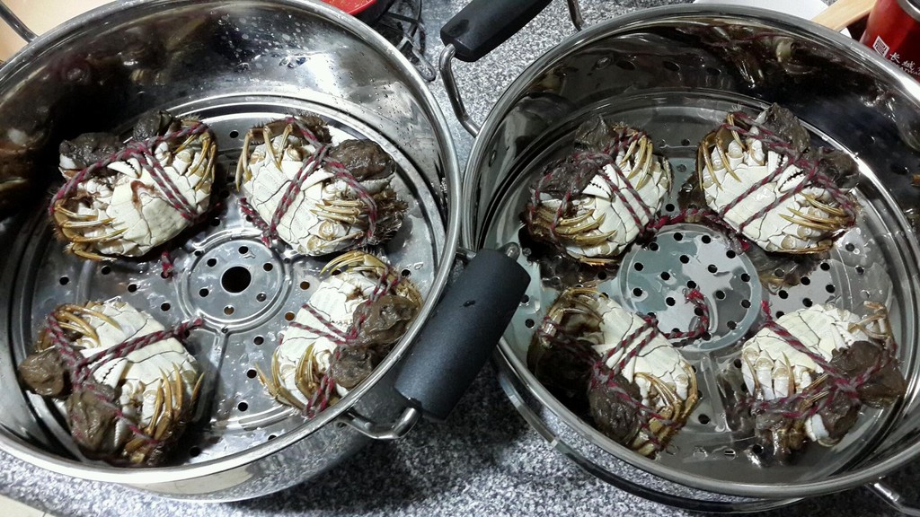 不锈钢蒸锅内正在蒸制两只绑着草绳的大闸蟹,蟹呈倒扣状,锅内蒸汽升腾,旁边放有生姜片和一瓶打开的花雕酒,展示传统清蒸做法 蒸制大闸蟹公蟹过程