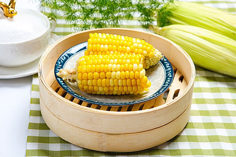 蒸锅复热糯玉米过程特写：不锈钢蒸格上整齐摆放4根解冻后的糯玉米，顶部覆盖湿润竹制蒸笼布，缕缕白汽升腾，背景虚化显示电子计时器显示“05:00”