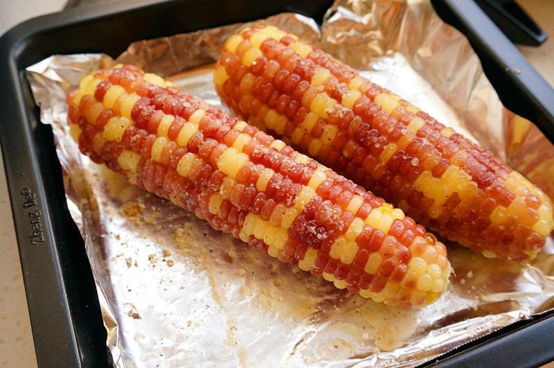 两根裹着金黄色蜂蜜黄油的糯玉米正在烤箱中烘烤,表面微微起泡,散发诱人的光泽,烤盘上有轻微焦痕,背景为厨房环境 烤箱中正在烘烤的糯玉米