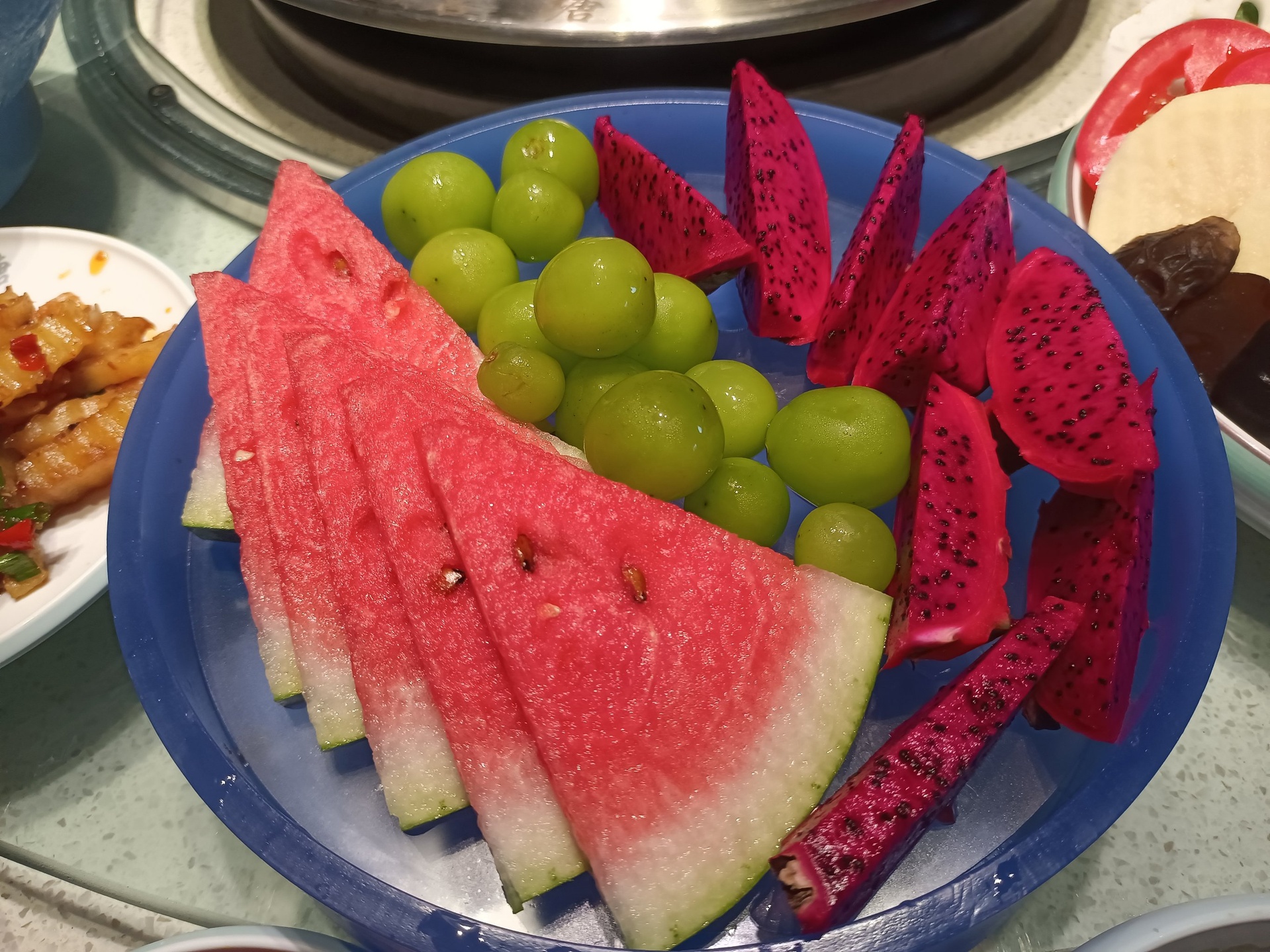 高清俯拍视角:西瓜青提拼盘完成态,突出果肉质地、颜色对比与天然光泽,白瓷盘衬托清爽感 西瓜青提拼盘成品特写:白瓷圆盘中央堆叠三层弧形西瓜块(深红瓤、薄翠皮),环绕放射状铺排整粒青提(青绿泛微霜,果蒂翠绿完整),顶部点缀三片薄荷叶与细碎柠檬皮丝,背景为浅木纹台面