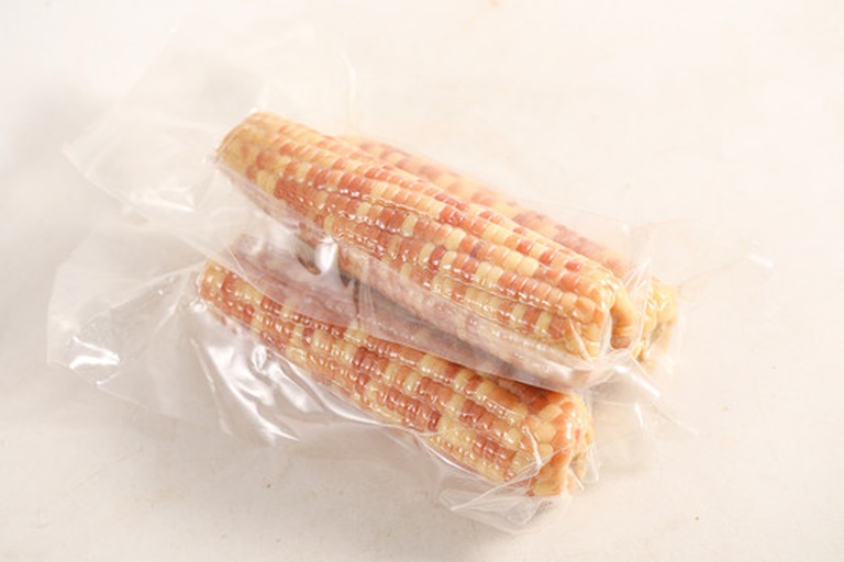 桌面上摆放着三种不同包装的拇指玉米产品:透明真空袋装、彩色礼盒装、以及开袋后摆盘在瓷盘中的蒸熟玉米,旁边配有叉子和柠檬片,展现其精致食用场景 电商包装与食用场景