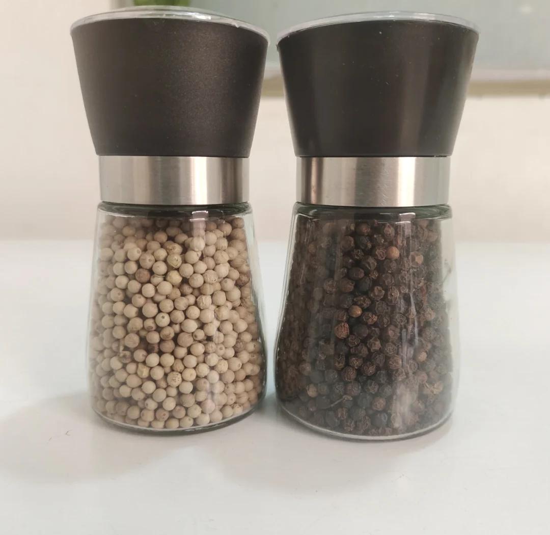 木质调料架上放置多个玻璃瓶,其中一个标有‘白胡椒’字样,瓶内为白色小颗粒。旁边配有手动研磨器,正在研磨出细粉,下方是一本打开的菜谱,页面写着‘白胡椒猪肚汤’的做法,展现家庭厨房中的实际应用场景 厨房中白胡椒储存与使用场景