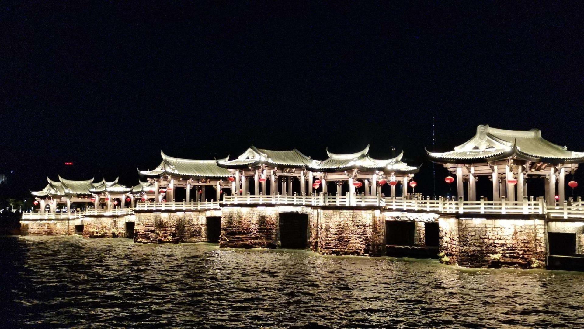 俯瞰视角下的混凝土防波堤延伸入海,夜晚灯光沿堤坝分布,几名垂钓者分散站立于不同位置,海面因灯光照射泛起涟漪,天空呈深蓝色,隐约可见星辰,整体氛围宁静而富有诗意 海边防波堤全景图