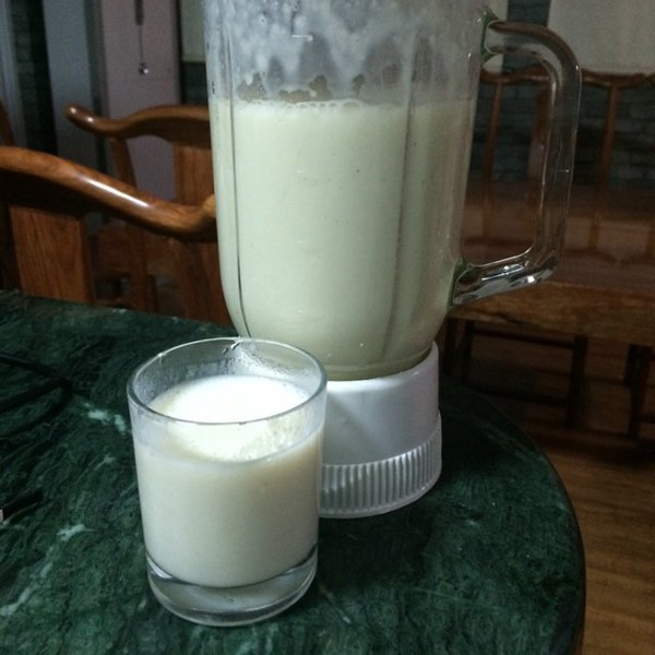 透明料理机正在搅打粉色液体,机器内可见奶油草莓块与牛奶混合旋转,旁边摆放着切好的草莓、牛奶盒、量杯和冰块,操作台整洁明亮,展现家庭自制饮品的专业流程 草莓牛奶制作过程图