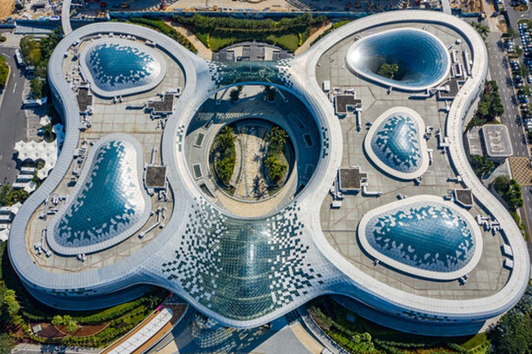 一座白色流线型的大型公共建筑,屋顶呈柔和的椭圆穹顶结构,阳光照射下呈现出类似蛋壳的弧度,周围绿植环绕,体现仿生学在建筑设计中的实际应用 现代建筑中仿生蛋形结构的应用实例