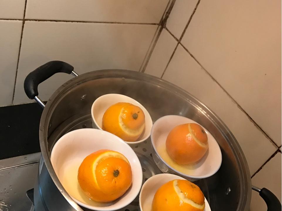 一个切开的冰糖橙放置在陶瓷碗中,顶部敞开,内部填入少许冰糖颗粒和几粒枸杞,旁边配有小勺和蒸锅,展示家庭蒸橙子的养生做法 冰糖橙蒸制过程