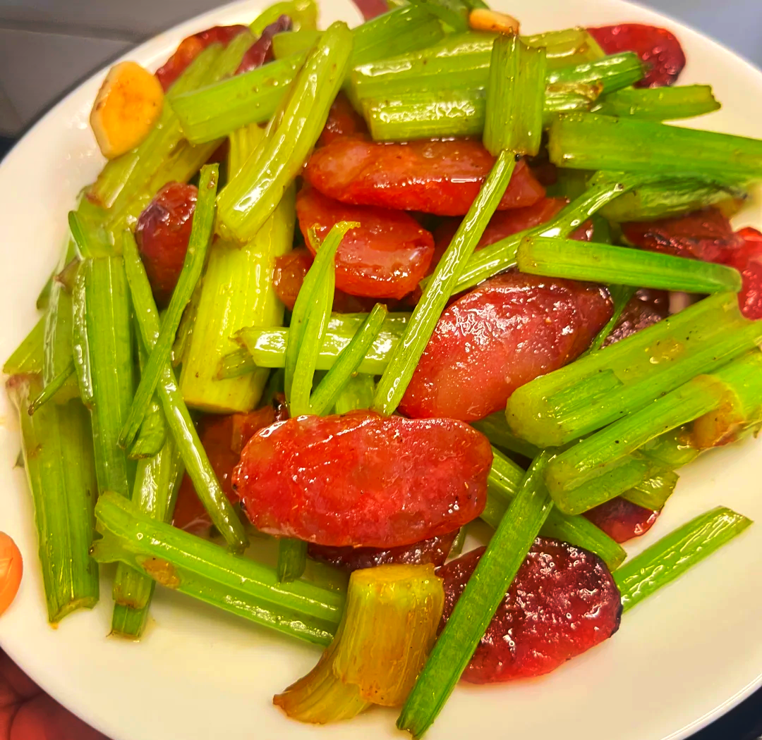 高清特写:左侧为新鲜翠绿、茎秆饱满带细绒毛的香芹(本芹),右侧为肥瘦均匀、表面微霜的广式腊肠切片;两者并置凸显颜色与质地对比,背景为木质砧板与不锈钢刀具 香芹与广式腊肠特写对比图