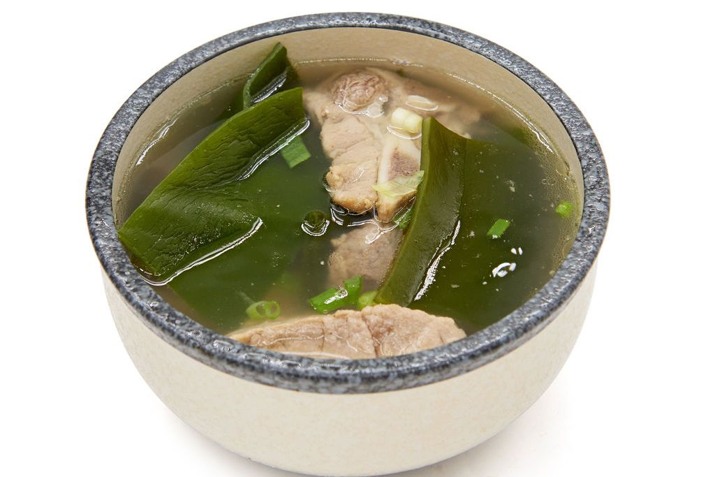 厨房灶台上正在熬煮的一锅乳白色海带排骨汤,汤面漂浮着绿色葱花和褐色海带片,旁边摆放着陶瓷汤碗和木勺,展现温馨家常饮食氛围 家庭烹饪海带汤场景