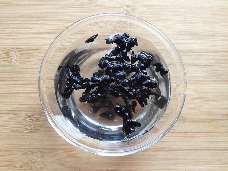 一个白色瓷碗中浸泡着膨胀中的黑木耳,另一口不锈钢锅中小火炖煮着加入胡萝卜和毛木耳的清汤,灶台旁摆放着剪刀、滤网和玻璃调料瓶,整体呈现家庭健康烹饪氛围 厨房中正在泡发与烹饪木耳的场景