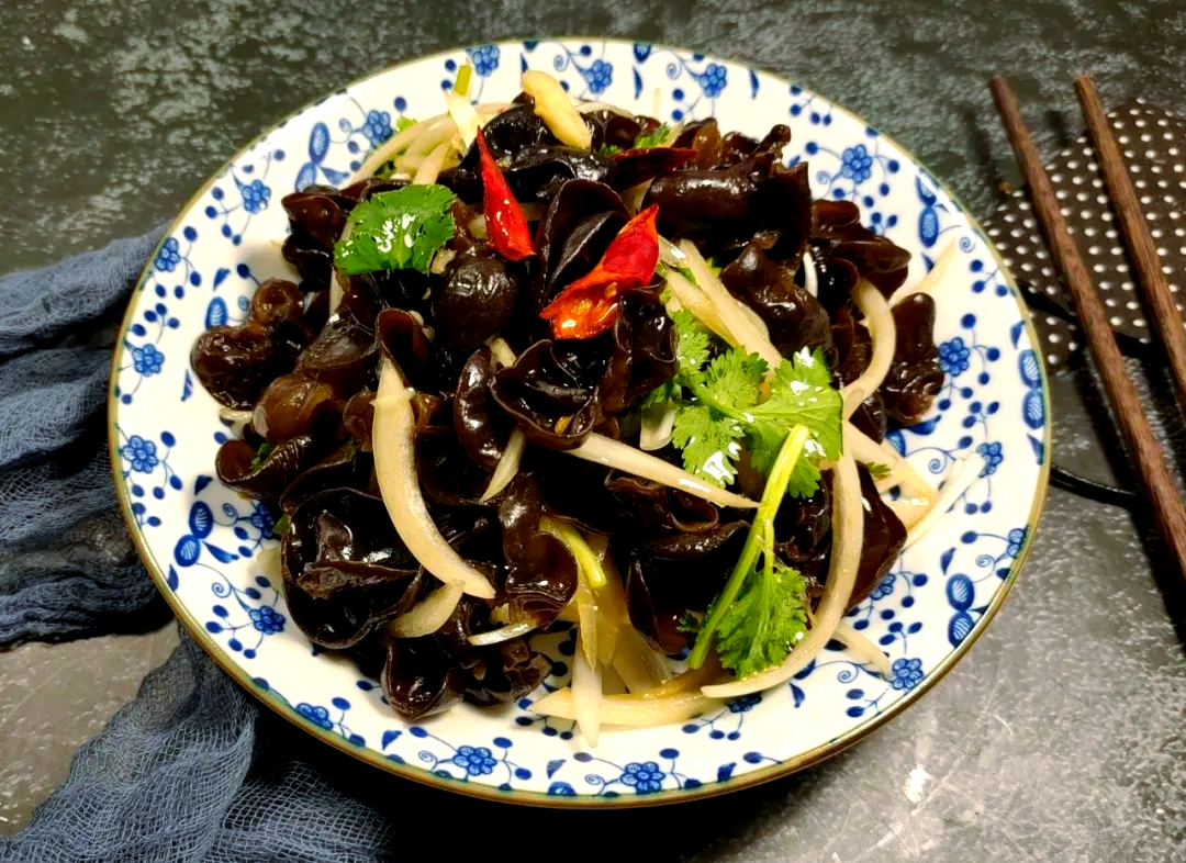 一张现代中式餐桌上的凉拌黑木耳菜品,黑色胶质感的木耳为主料,配有红色辣椒丝、绿色香菜段、蒜末和浅色调味汁,使用白色瓷盘盛放,背景虚化,突出食物色泽与质感 家庭餐桌上的一道凉拌黑木耳菜肴
