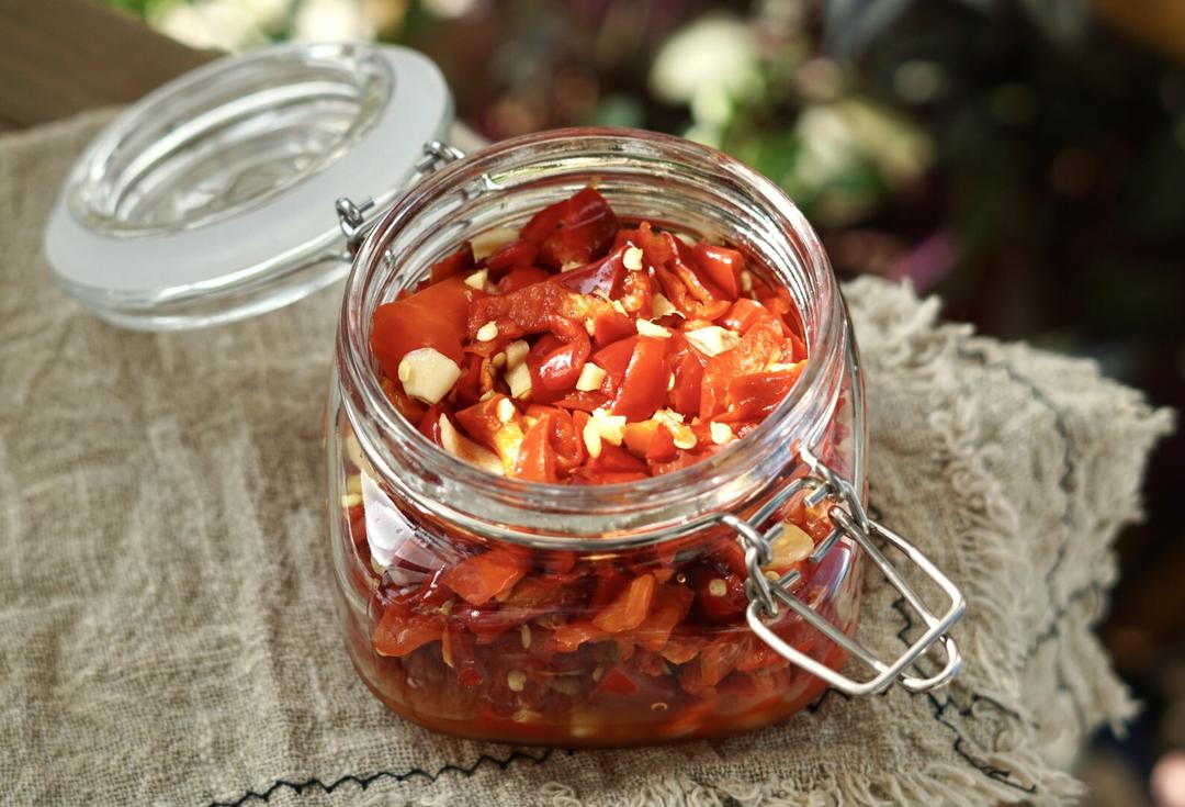 一瓶透明玻璃罐中盛满金红色的蒜米椒,可见清晰的蒜粒与辣椒段混合分布,表面覆盖一层透亮红油,标签手写‘自制蒜米椒’,放置在竹制餐垫上,旁边配有木勺和青菜 蒜米椒成品展示图